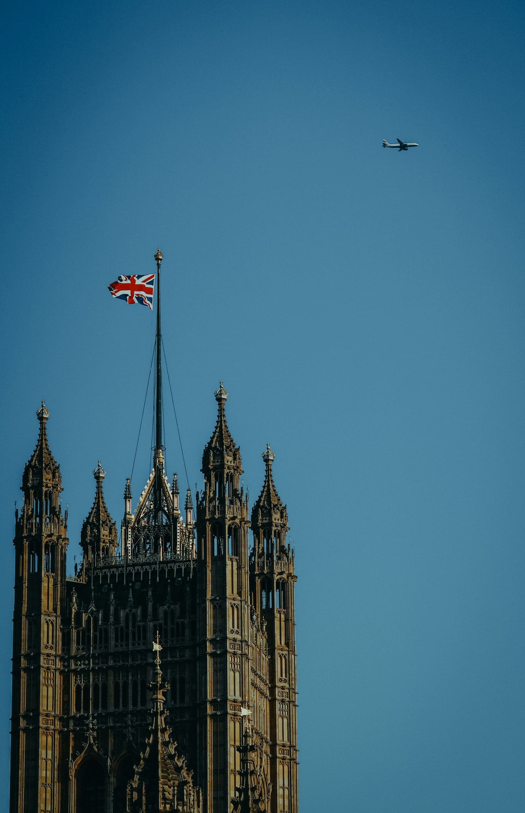 A Union Jack flying above the Houses of Parliament as an aircraft crosses the sky.