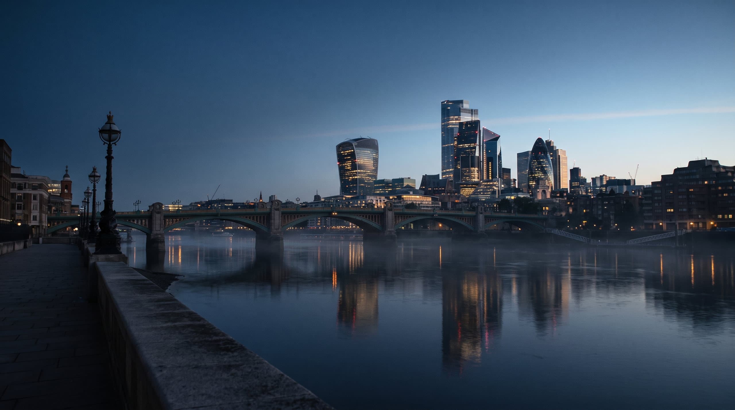 The City of London skyline at blue hour, seen across a still River Thames from an empty embankment.